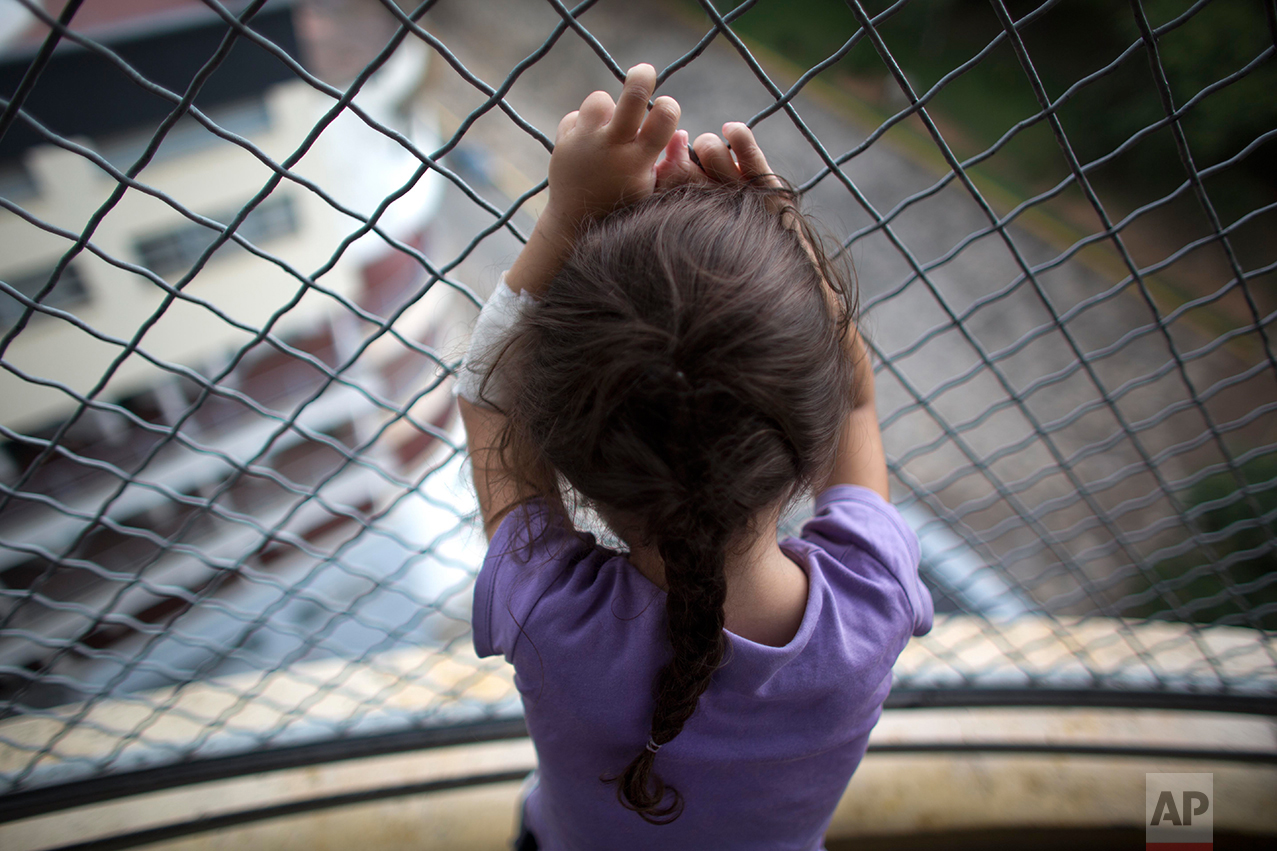 In this Aug. 23, 2016 photo, 3-year-old Ashley Pacheco hooks her fingers around the links of a fence as she gazes out from a hospital balcony, at University Hospital, in Caracas, Venezuela. This is the only place Ashley is able to breathe in fresh air. A scraped knee in mid-July led to a staph infection. There were not enough antibiotics to treat the infection, and so Ashley was kept in the public hospital for two months. (AP Photo/Ariana Cubillos)