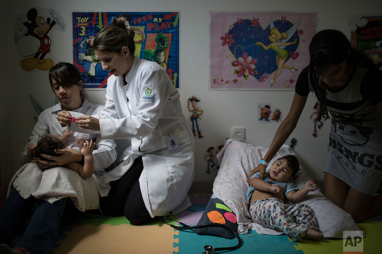In this Sept. 27, 2016 photo, physical therapist Camilla Costa, left, and speech therapist Amora Marins apply Kinesio Tape on Luiza as Victoria Leticia, right, holds her son Lucas Matheus, during a physical therapy session for babies born with microcephaly at the UPAE hospital in Caruaru, Pernambuco state, Brazil. A year after a spike in the number of newborns with microcephaly, doctors and researchers have seen many of the babies develop swallowing difficulties, epileptic seizures and vision and hearing problems. (AP Photo/Felipe Dana)