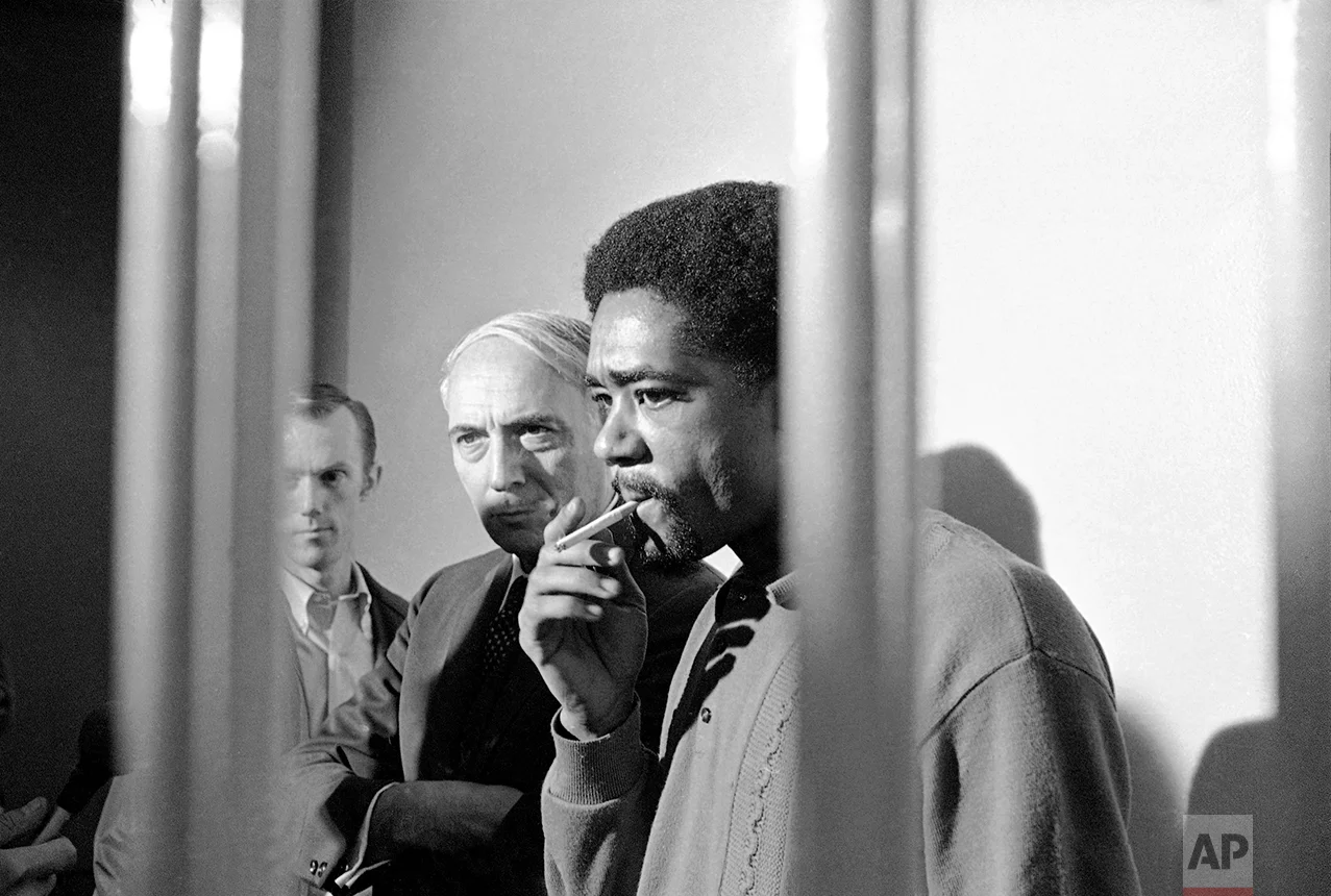 Standing in the visiting cell of the San Francisco City Prison, Bobby Seale, Black Panther leader, puffs on a cigarette during an informal news conference on Monday, Nov. 11, 1969 in San Francisco. Seale was returned to San Francisco from Chicago where he was sentenced to four years in jail on contempt of court charges. Seale was one of eight defendants charged with conspiracy in connection with the 1968 Democratic Convention riots in Chicago. Standing beside Seale is his attorney Charles Garry who could not represent him in Chicago because he had undergone surgery. (AP Photo)