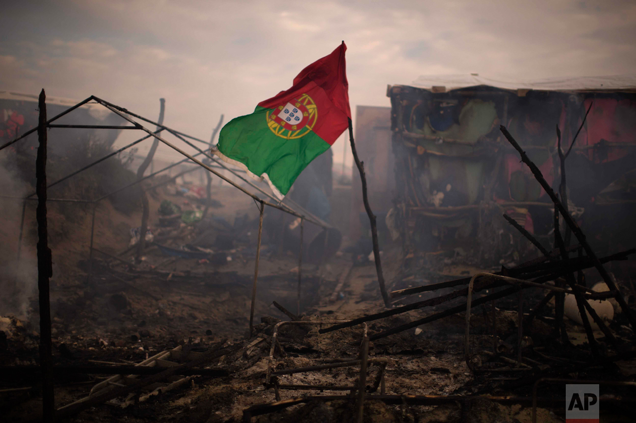 In this Wednesday, Oct. 26, 2016 photo, a flag of Portugal waves next to burning tents, abandoned by their owners at the makeshift migrant camp known as "the jungle" near Calais, northern France. (AP Photo/Emilio Morenatti)