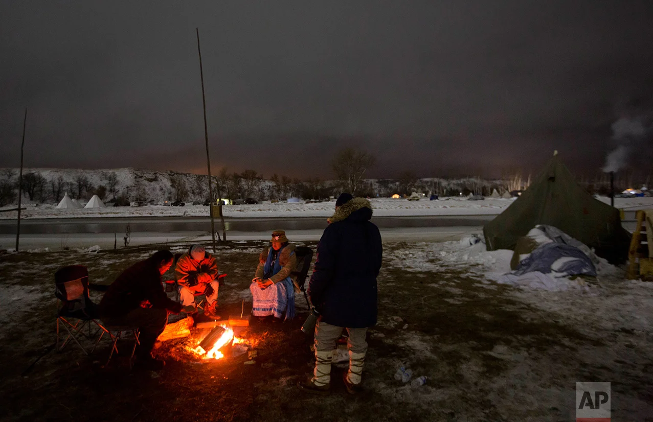 In this Thursday, Dec. 1, 2016 photo, campers sit around a fire along the Cannonball river at the Oceti Sakowin camp where people have gathered to protest the Dakota Access pipeline in Cannon Ball, N.D. So far, those fighting the Dakota Access pipeline have shrugged off the heavy snow, icy winds and frigid temperatures that have swirled around their encampment on the North Dakota grasslands. But if they defy next week's government deadline to abandon the camp, demonstrators know the real deep freeze lies ahead. (AP Photo/David Goldman)