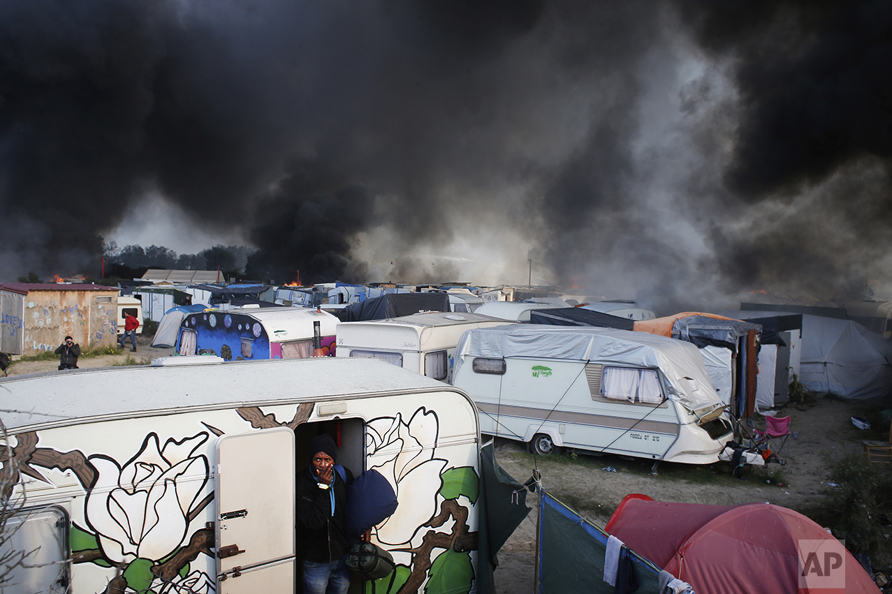  A man carrying his belongings reacts as he leaves a caravan, as smoke and flames rise from the tents after fires were started in the makeshift migrant camp known as "the jungle" near Calais, northern France, on Oct. 26, 2016. (AP Photo/Thibault Camu