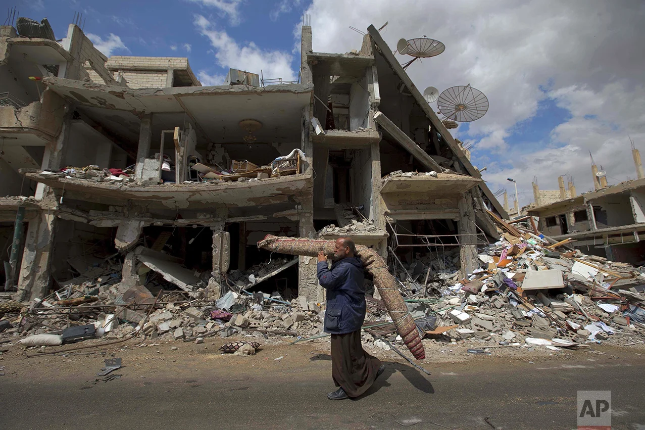 In this April 14, 2016 photo, a Syrian man carries a carpet through a devastated part of the town of Palmyra as families load their belongings onto buses in the central Homs province in Syria. Thousands of residents of this ancient town who fled Islamic State rule returned briefly to check on their homes and salvage what they could _ carpets, blankets, a refrigerator or a few family mementos. (AP Photo/Hassan Ammar)