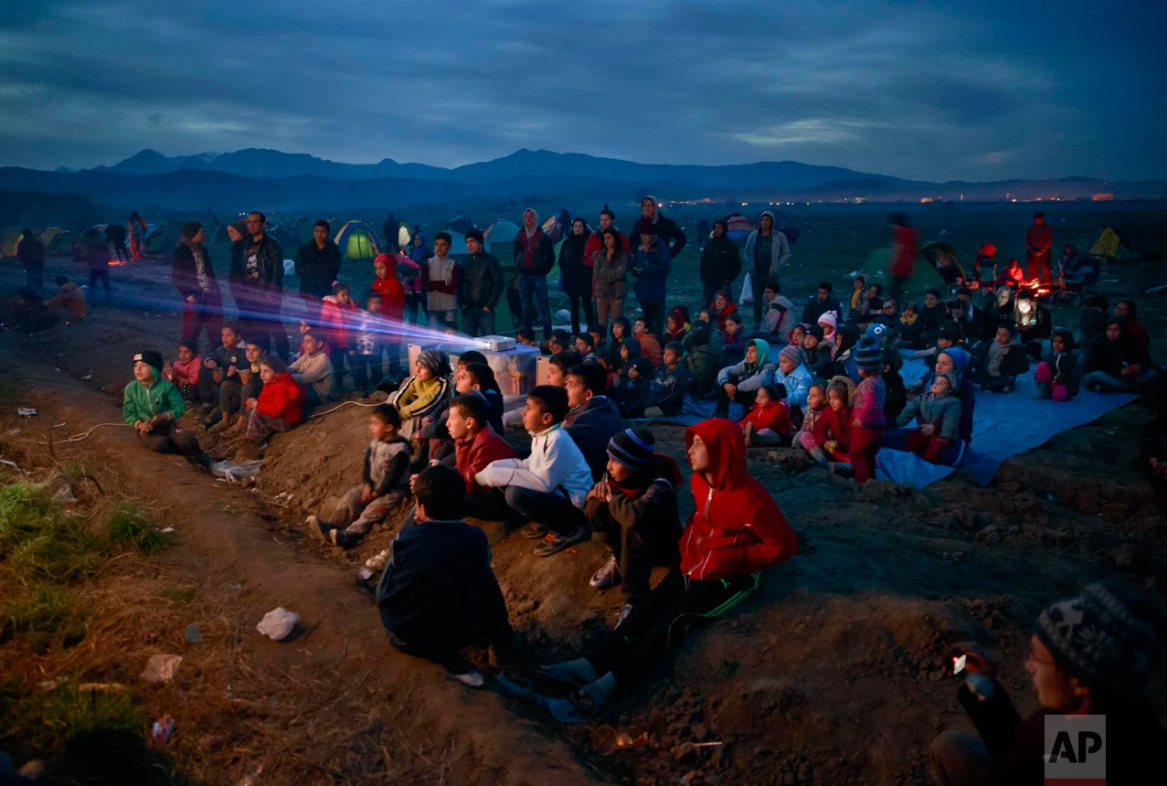 In this March 5, 2016 photo, children watch an animated movie in a field at the northern Greek border station of Idomeni. The Idomeni border crossing in the Greek region of Central Macedonia has become a bottleneck, where thousands of migrants are trapped as they try to find refuge and a better life in Europe. (AP Photo/Vadim Ghirda)