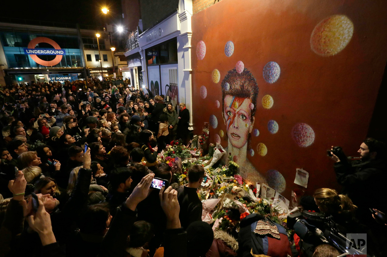  In this Monday, Jan. 11, 2016 photo, people gather next to tributes placed near a mural of British singer David Bowie by artist Jimmy C, in Brixton, south London. Bowie, the other-worldly musician who broke pop and rock boundaries with his creative 
