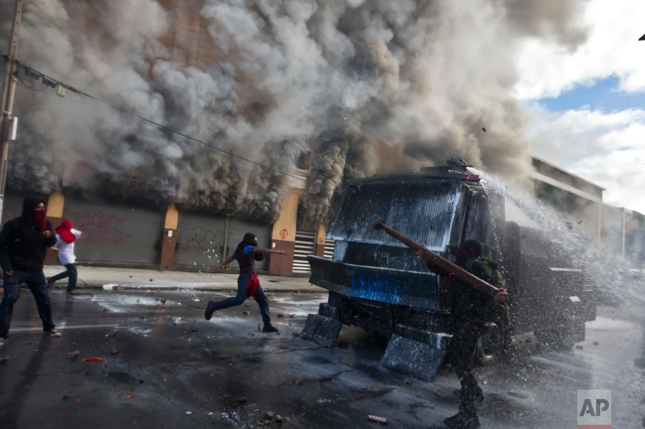 In this May 21, 2016 photo, masked protesters attack a police water canon in front of a burning pharmacy near Congress where President Michelle Bachelet was presenting the state-of-the-nation report, in Valparaiso, Chile. The anti-government protest began as a peaceful march but turned rough as some demonstrators threw rocks at police and gasoline bombs at buildings, resulting in the death of one man who reportedly died of asphyxiation. (AP Photo/Esteban Felix)