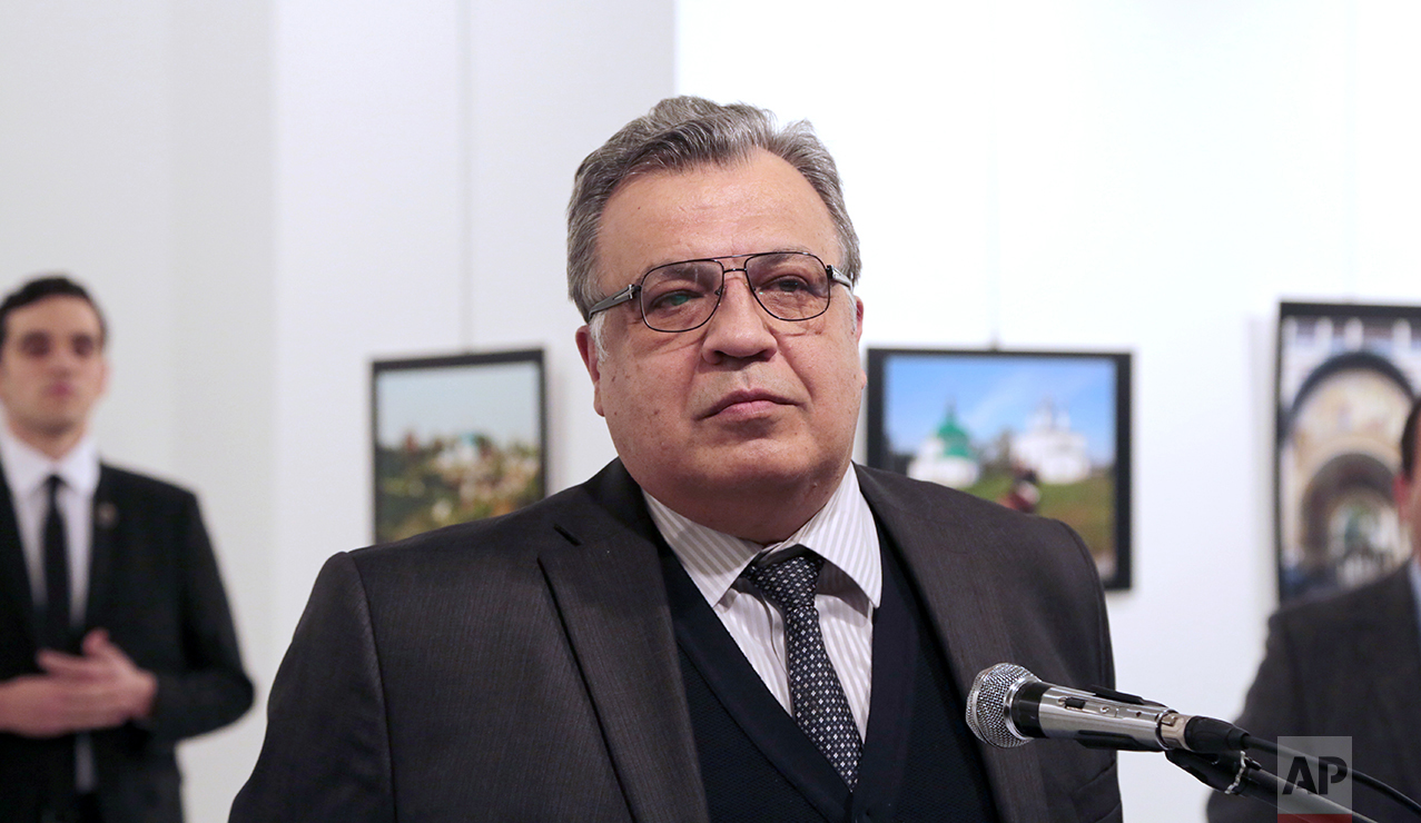 Andrei Karlov, the Russian Ambassador to Turkey, speaks at a photo exhibition in Ankara on Monday, Dec. 19, 2016, moments before a gunman opened fire on him. Karlov was rushed to a hospital after the attack and later died from his gunshot wounds. The gunman is seen at rear on the left. (AP Photo/Burhan Ozbilici)