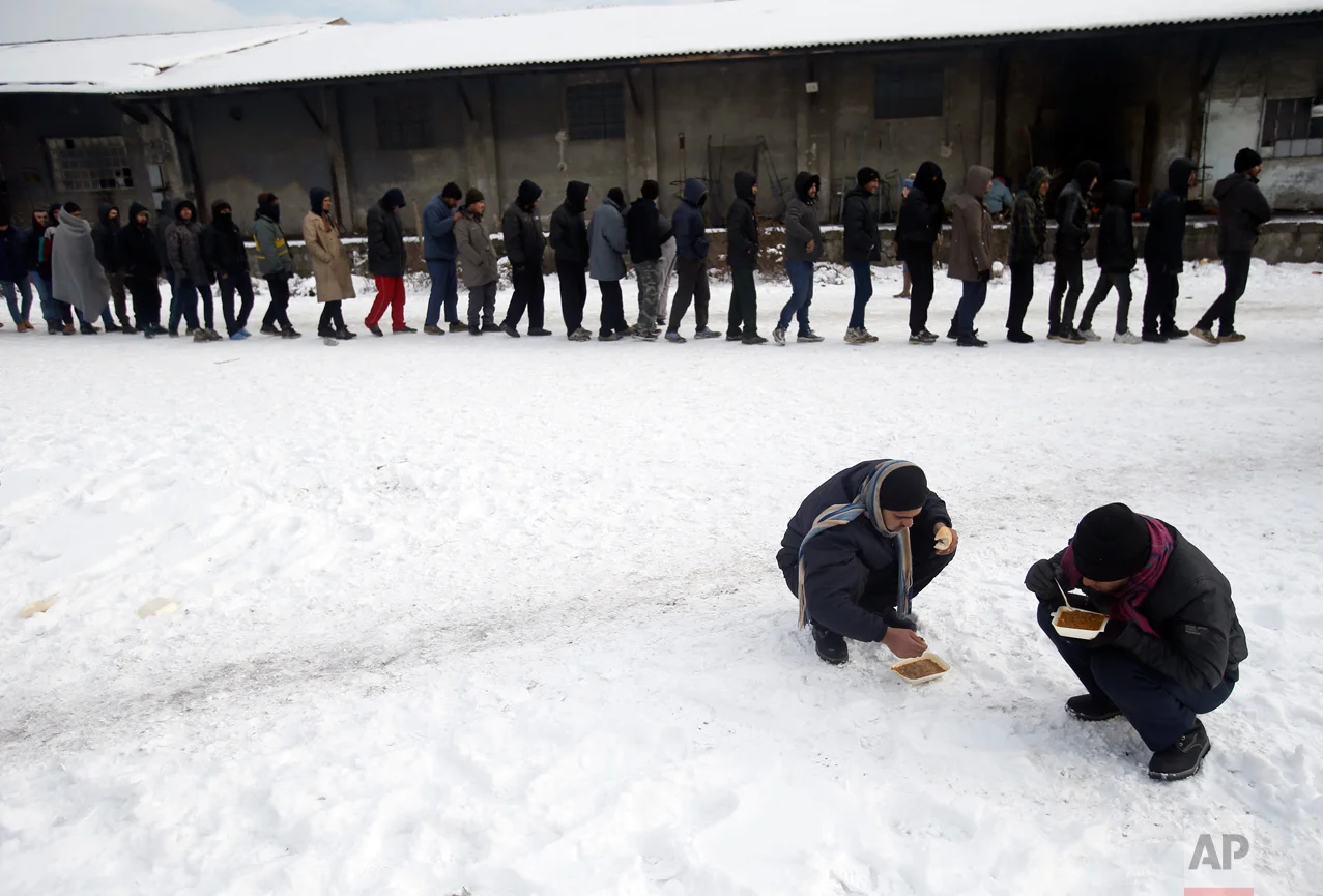In this Thursday, Jan. 12, 2017 photo, migrants squat in the snow as they eat warm meals distributed by aid groups as others queue for their portion outside a crumbling warehouse that has served as a makeshift shelter in Belgrade, Serbia. (AP Photo/Darko Vojinovic)