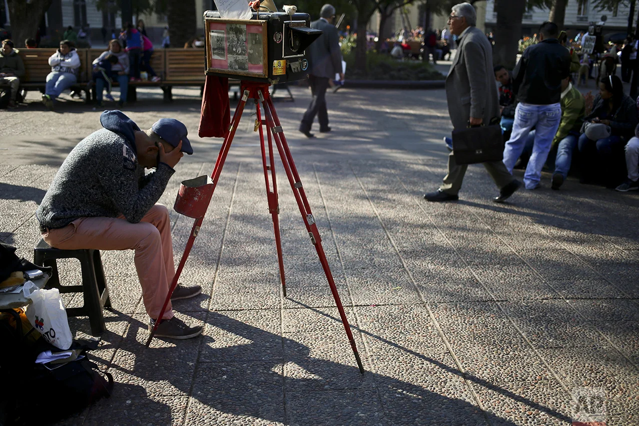 In this Sept. 1, 2016 photo, photographer Luis Maldonado waits for clients by his old wooden box camera in Plaza de Armas in Santiago, Chile. Clients are scarce, with days, even weeks, passing before someone asks him to create a portrait with the old-fashioned camera. (AP Photo/Esteban Felix)
