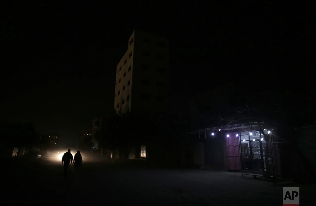 In this Monday, Jan. 16, 2017 photo, Palestinians walk on a dark street next to a grocery shop lit with a lamps powered by a battery during a power cut in Gaza City. (AP Photo/ Khalil Hamra)