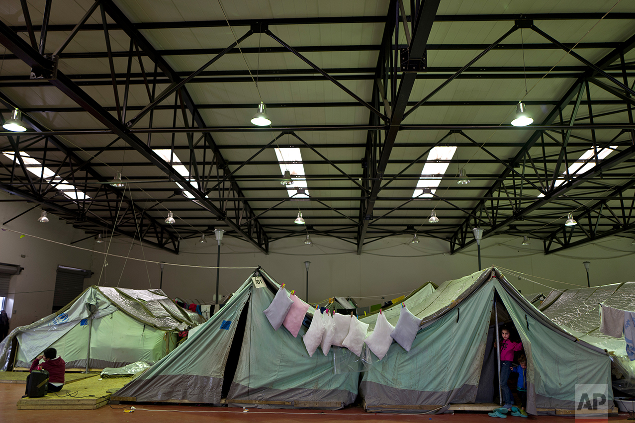 In this Thursday, Jan. 19, 2017 photo, a Syrian refugee boy, left, listens to music on speakers while a girl, right, peers with her younger sister through the entrance of their family's tent in Kalochori refugee camp on the outskirts of the northern Greek city of Thessaloniki. Many migrants living in this warehouse tent camp and another one nearby are feeling burned out. They try to keep busy as they dream of a better life in Western Europe and not let boredom or depression set in.(AP Photo/Muhammed Muheisen)