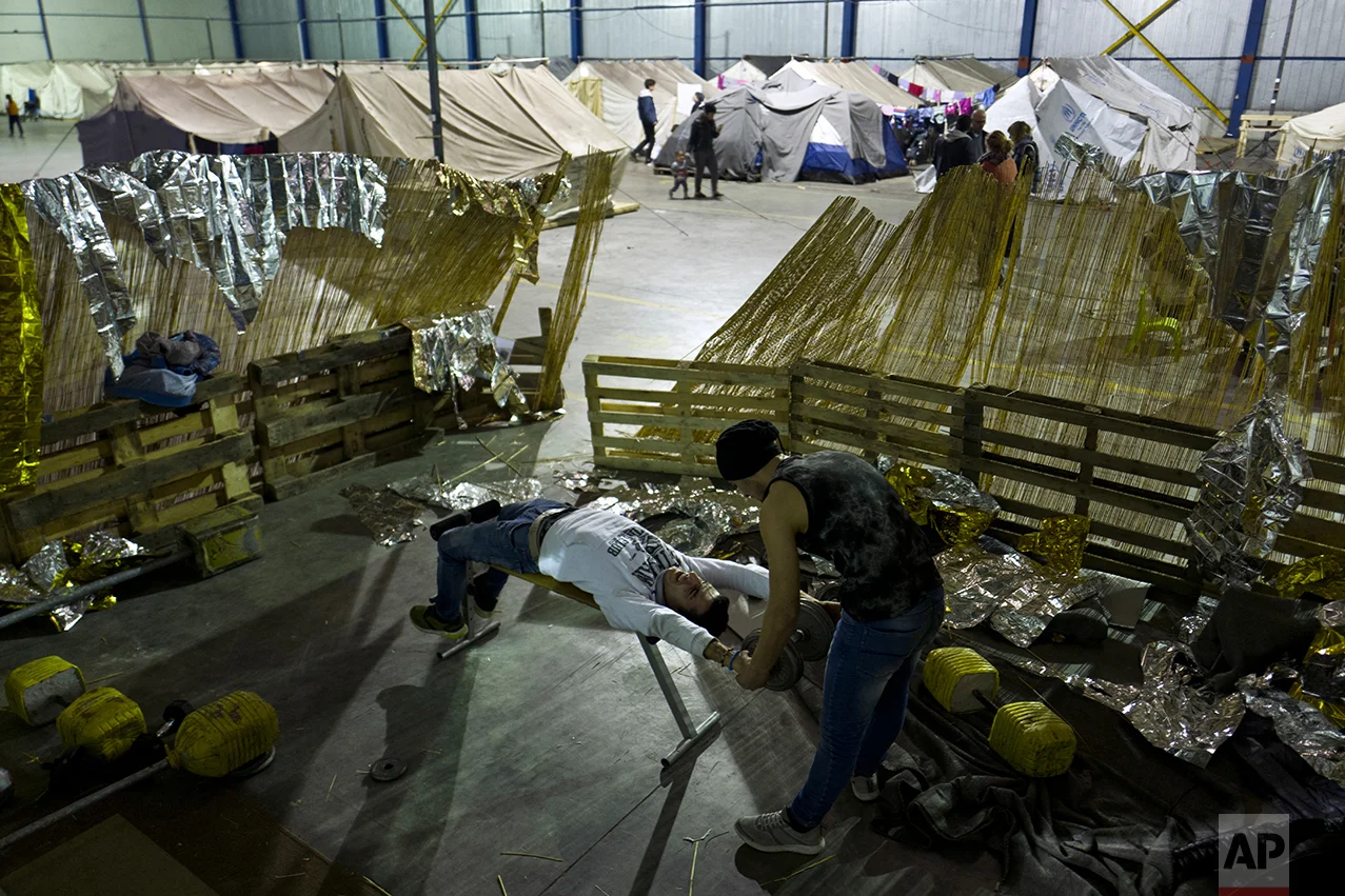 In this Thursday, Jan. 19, 2017 photo, a Syrian refugee boy, left, listens to music on speakers while a girl, right, peers with her younger sister through the entrance of their family's tent in Kalochori refugee camp on the outskirts of the northern Greek city of Thessaloniki. Many migrants living in this warehouse tent camp and another one nearby are feeling burned out. They try to keep busy as they dream of a better life in Western Europe and not let boredom or depression set in.(AP Photo/Muhammed Muheisen)