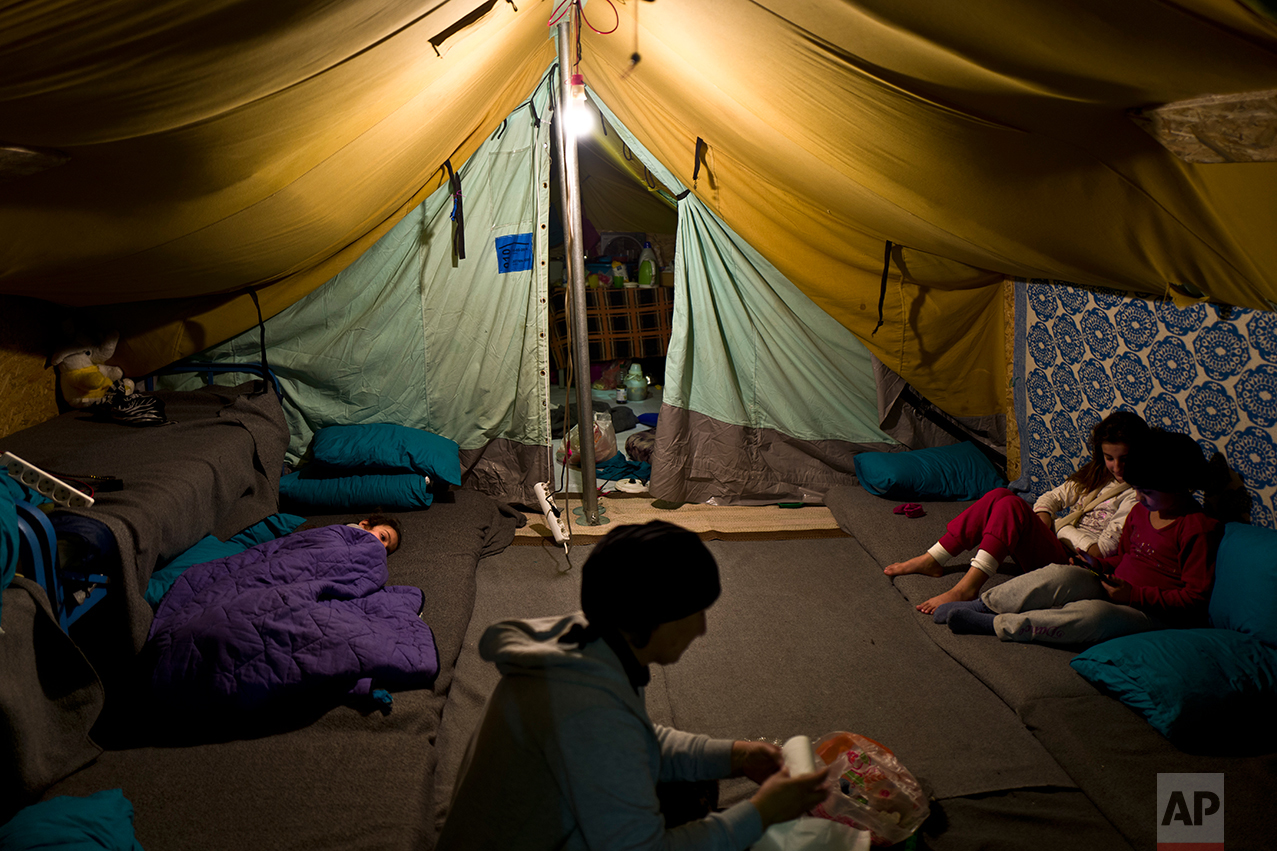In this Tuesday, Jan. 17, 2017 photo, a Syrian refugee woman works inside her tent while her daughter left lies on the ground and her other daughter play with her friends, in Kalochori refugee camp on the outskirts of the northern Greek city of Thessaloniki. (AP Photo/Muhammed Muheisen)