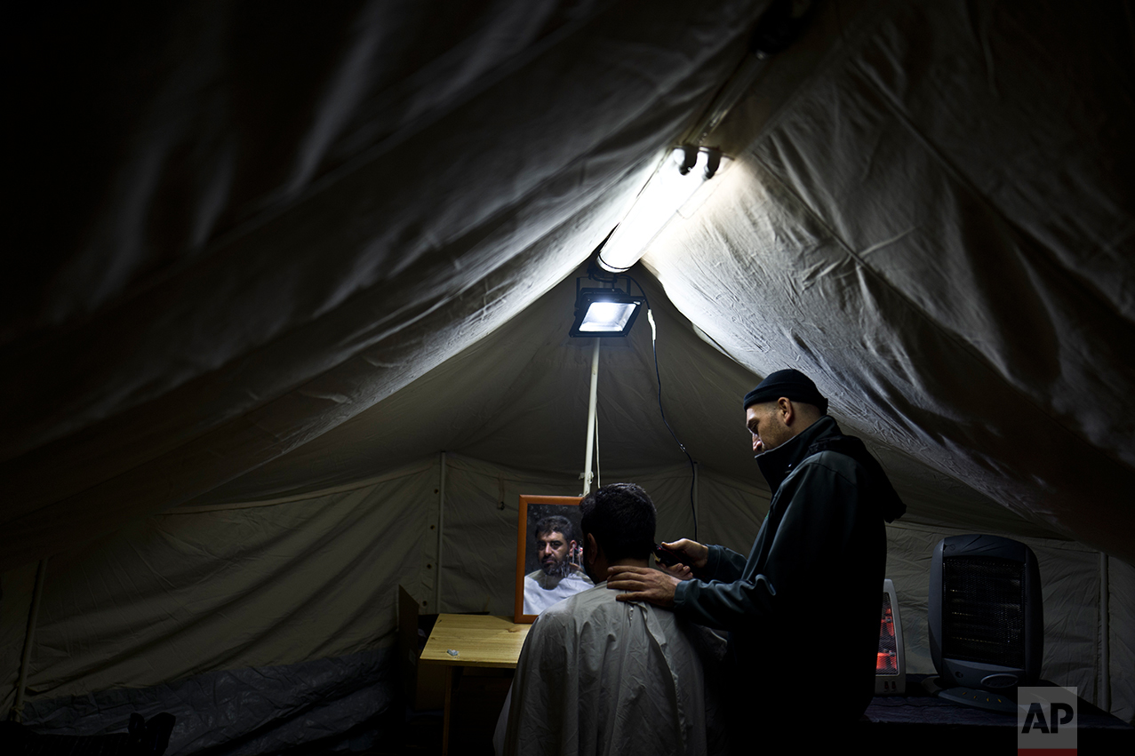 In this Wednesday, Jan. 18, 2017 photo, Syrian refugee Rewank Khalil, 30, shaves the hair of his friend Haitham Omar, 35, at a tent in Frakapor refugee camp on the outskirts of the northern Greek city of Thessaloniki. Because of a harsh winter and bitter cold, many migrants rarely venture out and away from the monotony and fluorescent lights hanging overhead. (AP Photo/Muhammed Muheisen)