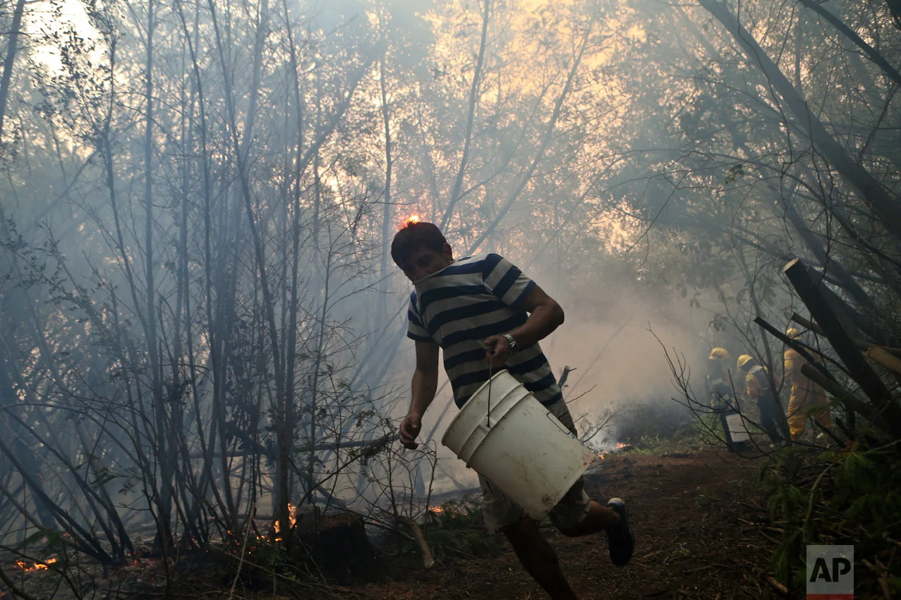 In this Saturday, Jan. 28, 2017 photo, a resident helping firefighters runs with an empty water bucket as they fight wildfires in Paso El Leon, in Concepcion, Chile. Residents of some communities have been battling the fires themselves, without any protective gear and often using just branches or bottles of water in a frantic effort to save their homes, pasture and livestock. (AP Photo/Esteban Felix)