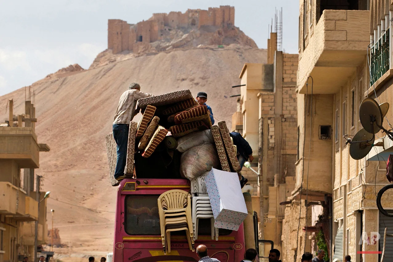 In this picture taken Thursday, April 14, 2016, the Palmyra citadel is seen in the background as Syrian families load their belongings onto a bus in the town of Palmyra in the central Homs province, Syria. Thousands of residents of this ancient town who fled Islamic State rule are returning briefly to check on their homes and salvage what they can - some carpets, blankets, a fridge or a few family mementos. (AP Photo/Hassan Ammar)