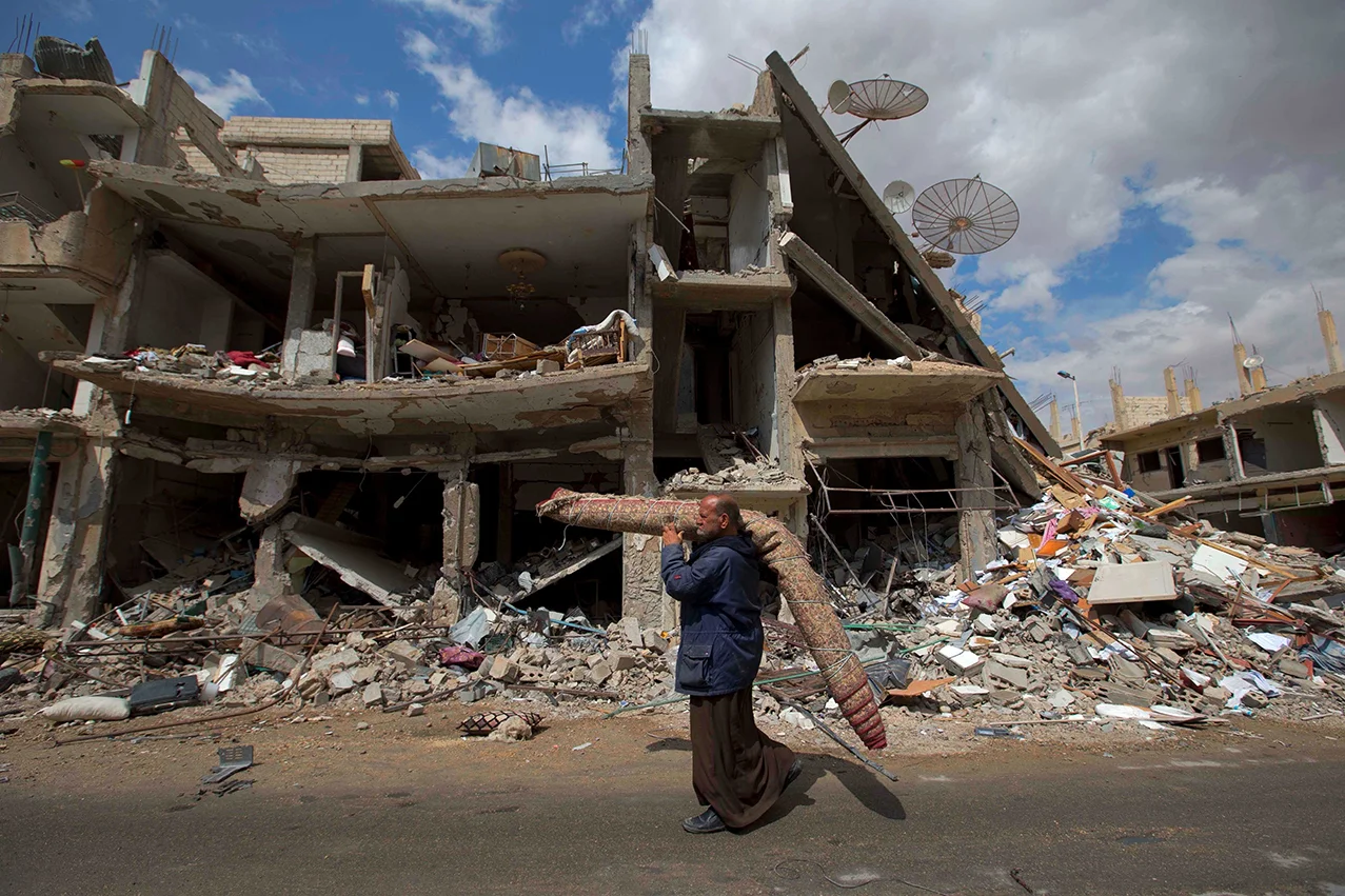 In this picture taken Thursday, April 14, 2016, a Syrian man carries a carpet as walk through a devastated part of the town of Palmyra as families load their belongings onto a bus in the central Homs province, Syria. Thousands of residents of this ancient town who fled Islamic State rule are returning briefly to check on their homes and salvage what they can – some carpets, blankets, a fridge or a few family mementos. (AP Photo/Hassan Ammar)