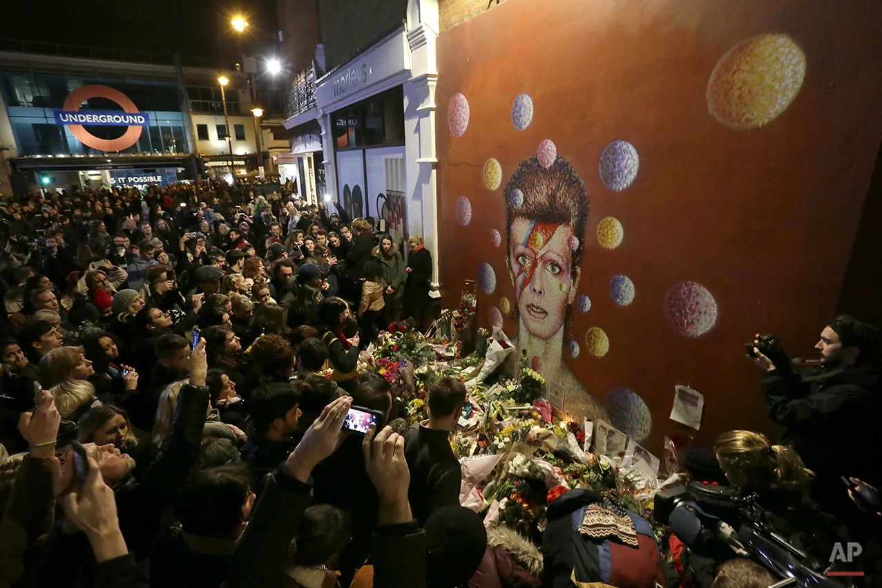 People gather next to tributes placed near a mural of British singer David Bowie by artist Jimmy C, in Brixton, south London, Monday, Jan. 11, 2016. (AP Photo/Tim Ireland)