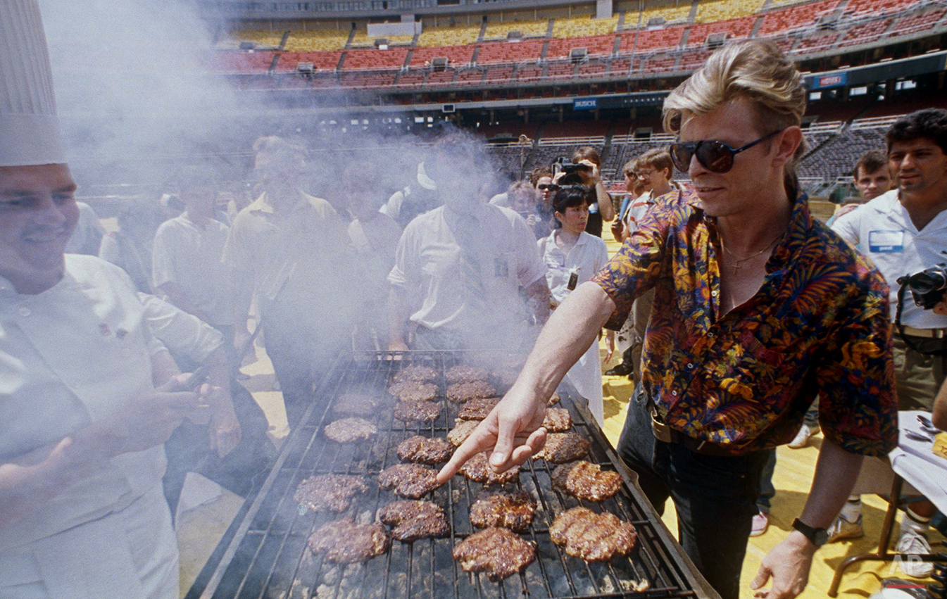 Rocker David Bowie, right, points to the hamburger that he wants for lunch during a barbecue for his crew at Veterans Stadium in Philadelphia, July 27, 1987. (AP Photo/Charles Krupa)