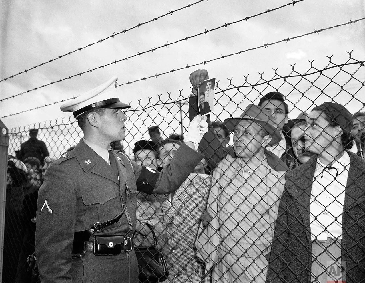 In this Nov. 21, 1951 photo, Bill Vetesy of Colonia, N.J., holds a picture of his mother and brother as he asks a military police officer inside the fence at Camp Kilmer, N.J., if they are among the 60 Hungarian refugees who arrived at the camp. The first plane load of refugees arrived at nearby McGuire Air Force Base from Vienna earlier in the day. The refugees will be housed at Camp Kilmer until quarters in private homes are made available to them in the United States. (AP Photo/Anthoy Camerano)
