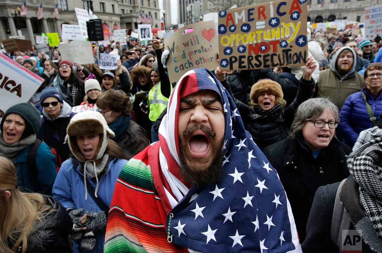 Izzy Berdan, center, wears a U.S. flag as he chants slogans with other demonstrators during a rally against President Donald Trump's order that restricts travel to the United States, Sunday, Jan. 29, 2017, in Boston. Trump signed an executive order Friday, Jan. 27, 2017 that bans legal U.S. residents and visa-holders from seven Muslim-majority nations from entering the U.S. for 90 days and puts an indefinite hold on a program resettling Syrian refugees. (AP Photo/Steven Senne)