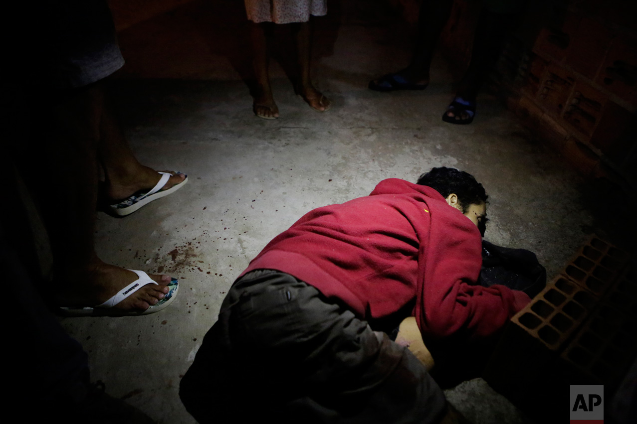 In this Thursday, Feb 9, 2017 photo, Nelson Eduardo Conclaves lies in a pool of blood as a neighbor stands over him in Vitoria, Espirito Santo state, Brazil. According to his mother Erlita Pereira Goncalves, her 30-year-old son was shot dead by attackers who broke into their home and killed him in front of her. (AP Photo/Diego Herculano)