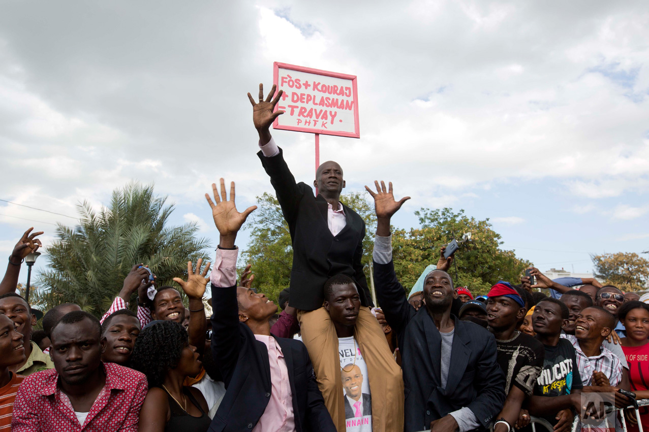 A supporter of Haiti's President Jovenel Moise impersonates the new president, with a sign behind him that reads in Creole "Energy plus encouragement plus movement plus work" during Moise's inauguration in Port-au-Prince, Haiti, Tuesday, Feb. 7, 2017. Moise was sworn-in as president for the next five years after a bruising two-year election cycle, inheriting a struggling economy and a deeply divided society. (AP Photo/Dieu Nalio Chery)