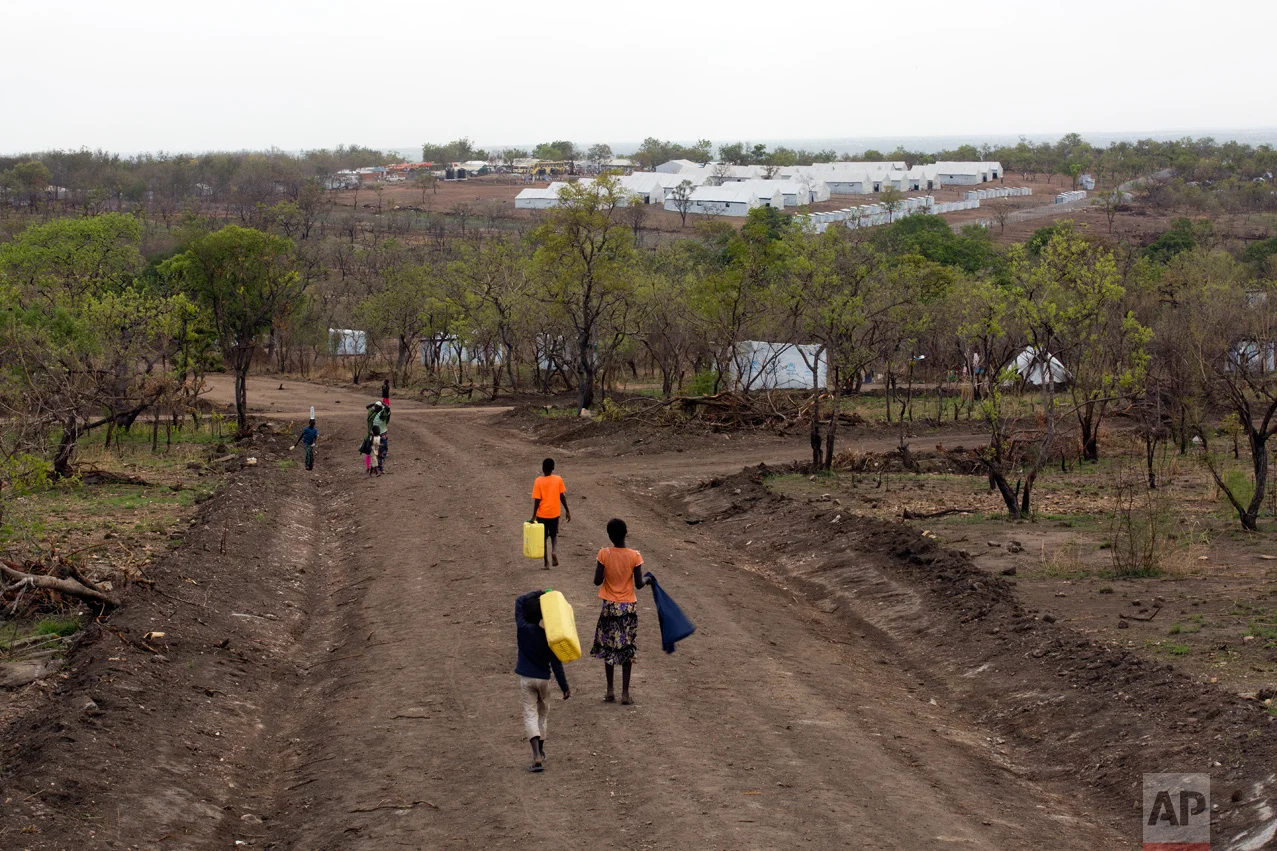 In this Thursday, April 6, 2017 photo, South Sudanese children carry water jugs down a road in the new Imvepi refugee settlement in northern Uganda. In the background are the communal tents for the processing center. Imvepi is growing at a rate of over 2,000 refugees each day. (AP Photo/Jerome Delay)