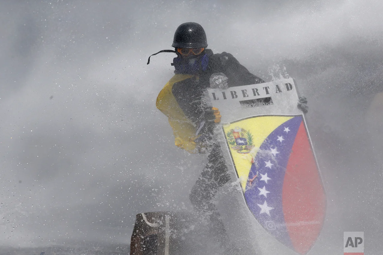 In this May 10, 2017 photo, amid a cloud of tear gas, an anti-government protester wearing a helmet, googles, gas mask and gloves holds a shield with the Spanish word "Freedom," to protect himself from a water cannon fired by security forces in Caracas, Venezuela. The government's response to the demonstrations has drawn international condemnation, with U.S. Secretary of State Rex Tillerson expressing concern in April that Maduro is "not allowing the opposition to have their voices heard." (AP Photo/Ariana Cubillos)