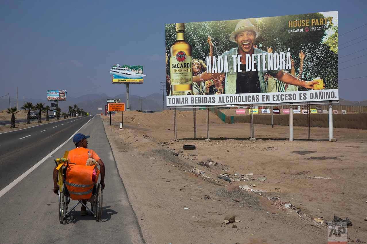  In this May 5, 2017 photo, Jose Suarez, from Cucuta, Colombia, moves his wheelchair, decorated with a stuffed animal, along the Pan American Highway lined with billboards advertising rum, lottery and mixed nuts, on the south side of Lima, Peru. Suar