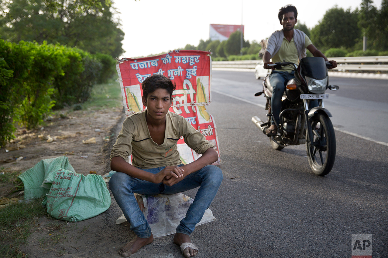  In this Tuesday, June 6, 2017 photo, an Indian boy who sells corn along a busy expressway poses for a photo next to his corn stall as a man on a motorcycle patrols to ensure business runs smoothly in Noida, India. Every 100 meters (330 feet) or so t