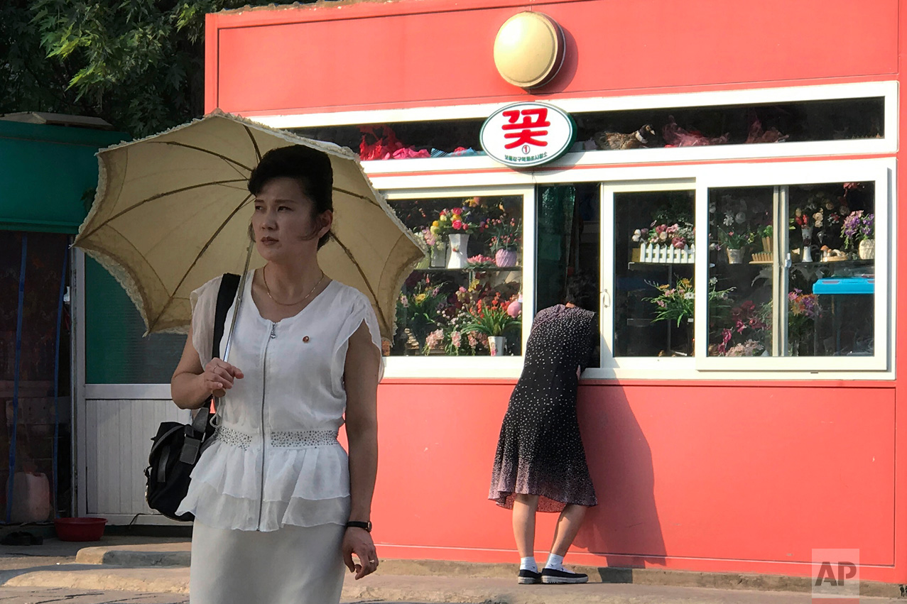 A woman looks into the window of a kiosk selling gifts and flowers in Pyongyang, North Korea, as a pedestrian waits to cross the street on Friday, June 16, 2017. Kiosks like these are a common sight around the city, some also selling food and drinks. (AP Photo/Wong Maye-E)