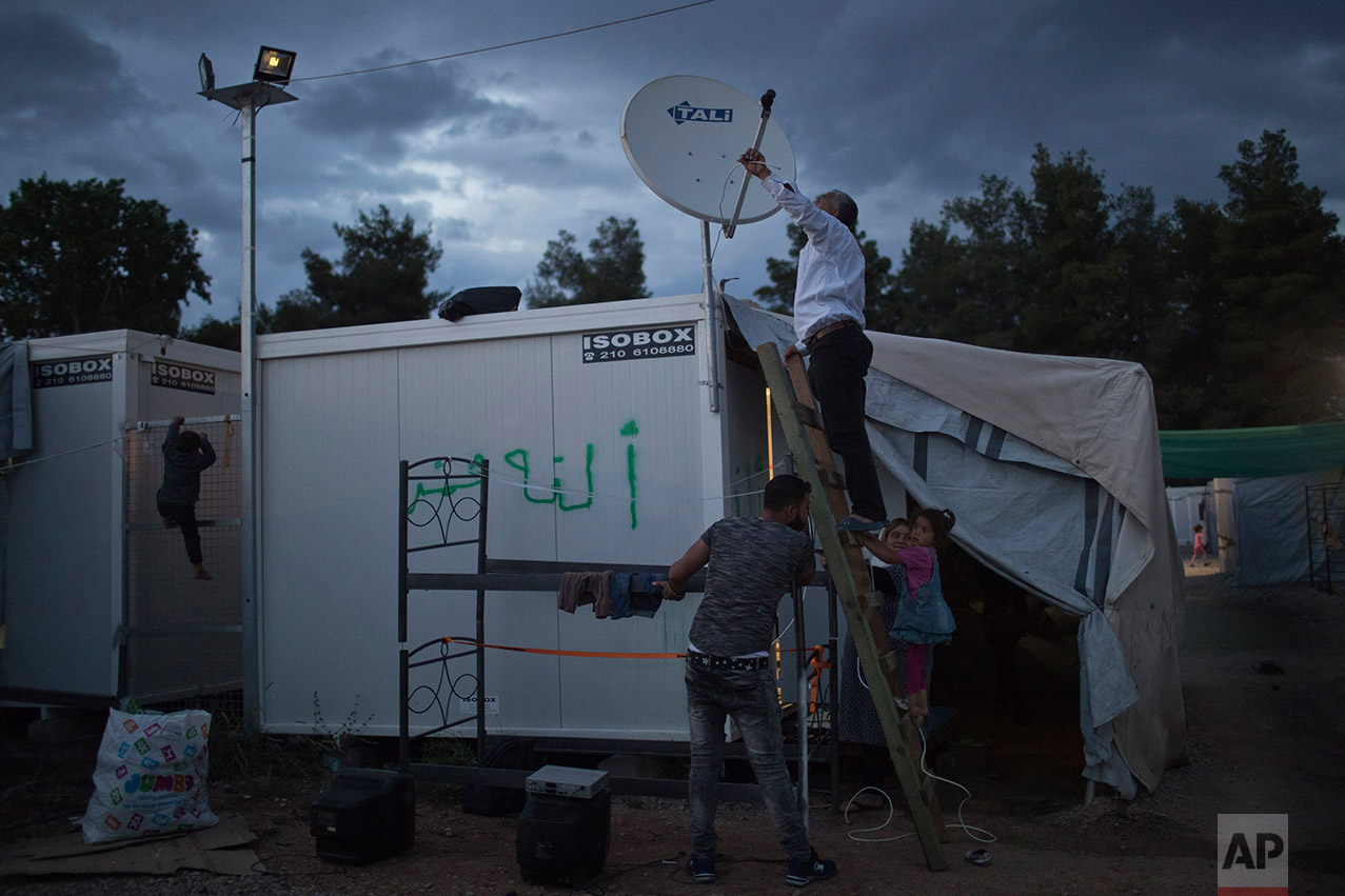  In this May 26, 2017 photo, refugees try to connect a cable to a satellite dish as a child climbs on an iron fence at the refugee camp of Ritsona about 86 kilometers (53 miles) north of Athens. On the mainland, migrant children receive after-hours c