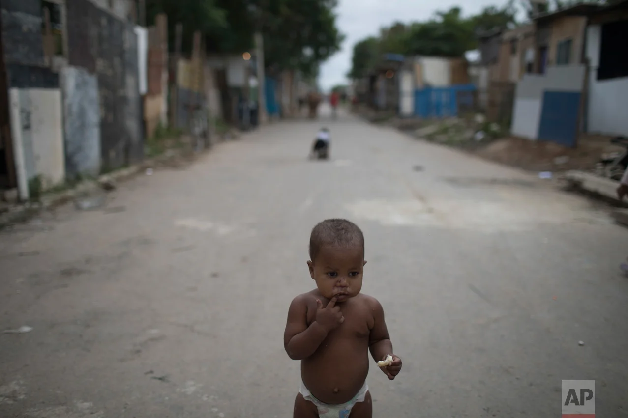 In this June 10, 2017 photo, a baby with a piece of bread stands outside his home under the watch of his mother in the neighborhood "Cantinho do Ceu" which means in Portuguese "Heaven's corner," one of the poorest areas of the City of God slum of Rio de Janeiro, Brazil. (AP Photo/Leo Correa)