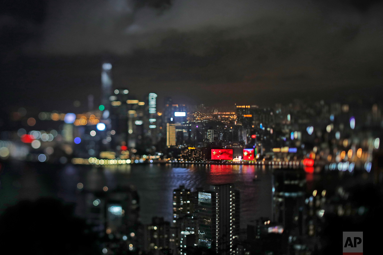 In this June 24, 2017, photo made with a tilt-shift lens, a Chinese national flag appears on a screen to mark the 20th handover anniversary at Victoria Habor in Hong Kong. Hong Kong's famous waterfront retains its British colonial era name and was the scene of Britain's farewell from its colony. Since Beijing took control of Hong Kong in 1997, China's rising influence – and Britain's waning profile – are impossible to ignore. (AP Photo/Vincent Yu)