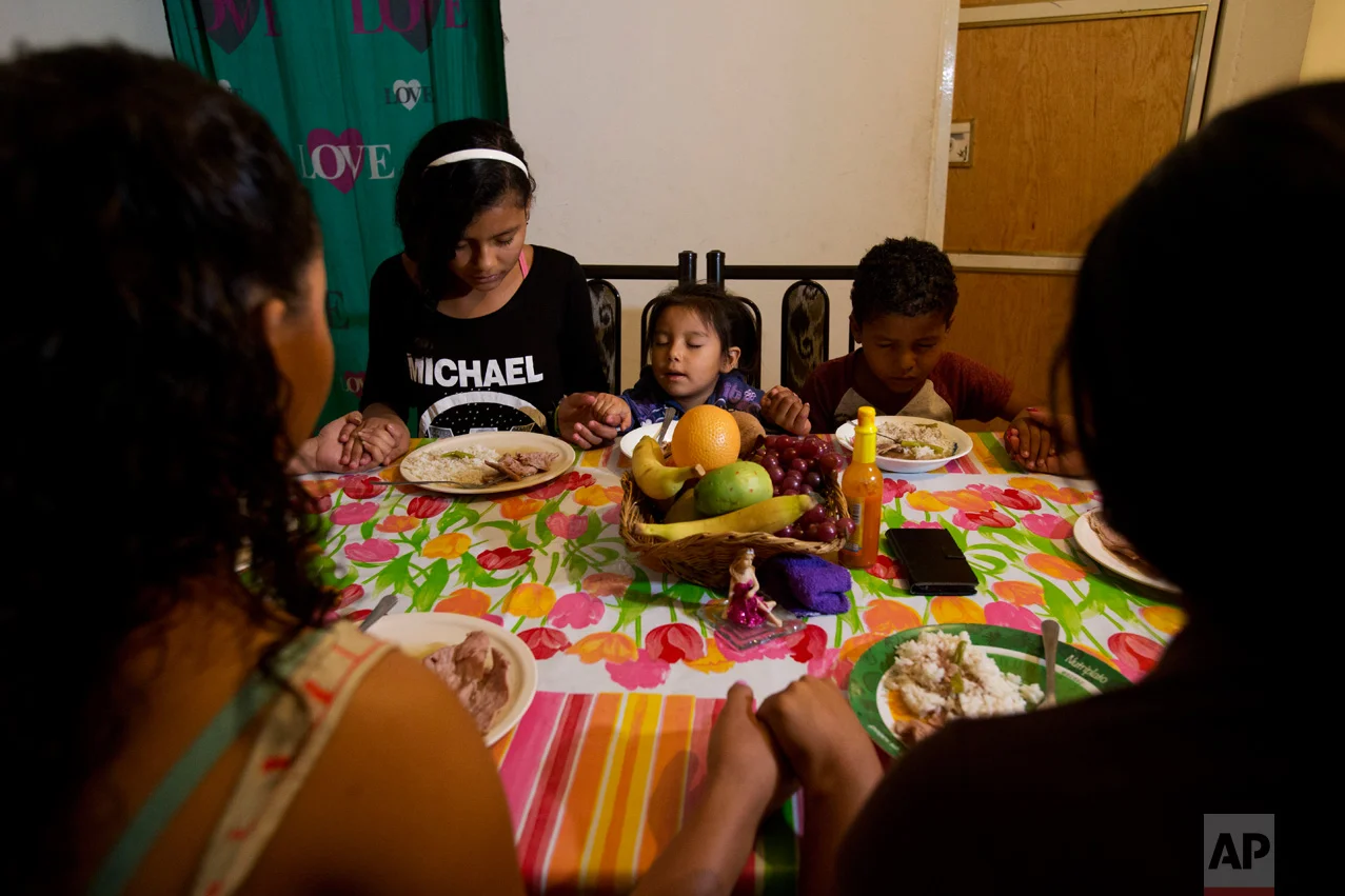 In this June 27, 2017 photo, three-year-old Carlita Perez, center, from El Salvador, says grace along with, from left, Honduran siblings Laura, Ruth, and Josue Funez Cruz, and their cousin Emma Karina Cruz, as they sit down to dinner in the two-bedroom apartment the two families share in the Iztapalapa district of Mexico City. (AP Photo/Rebecca Blackwell)