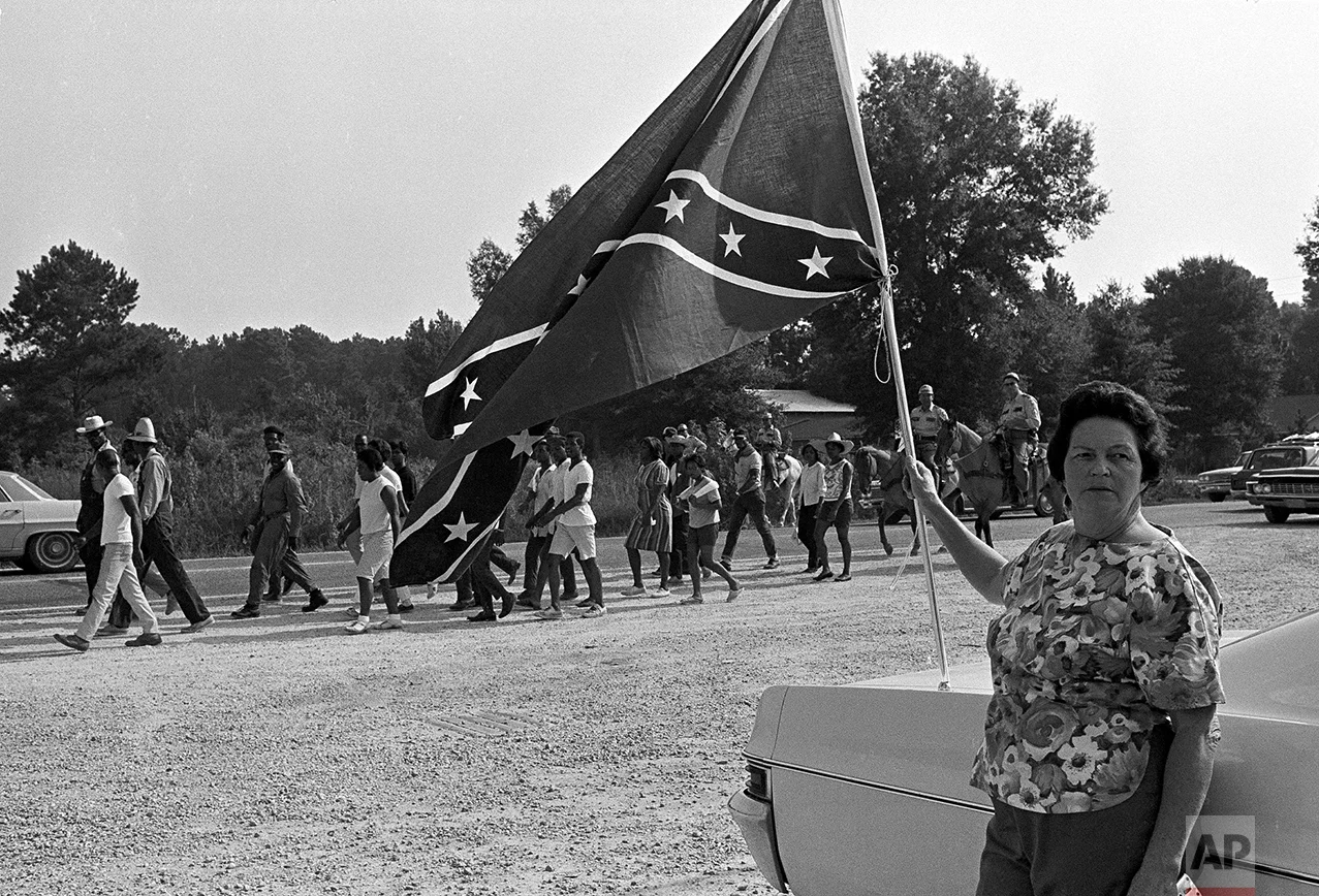 A white woman stands in protest by a confederate flag fluttering in the breeze as a group of 20 civil rights marchers pass through Hammond, La., Aug. 14, 1967. Racial shooting erupted last night in Hammond leaving five white men wounded. Twelve African-Americans were arrested in the shooting. The marchers, who camped at Hammond last night, are en route to Baton Rouge, La., to present a list of demands to the governor. (AP Photo/Jack Thornell)