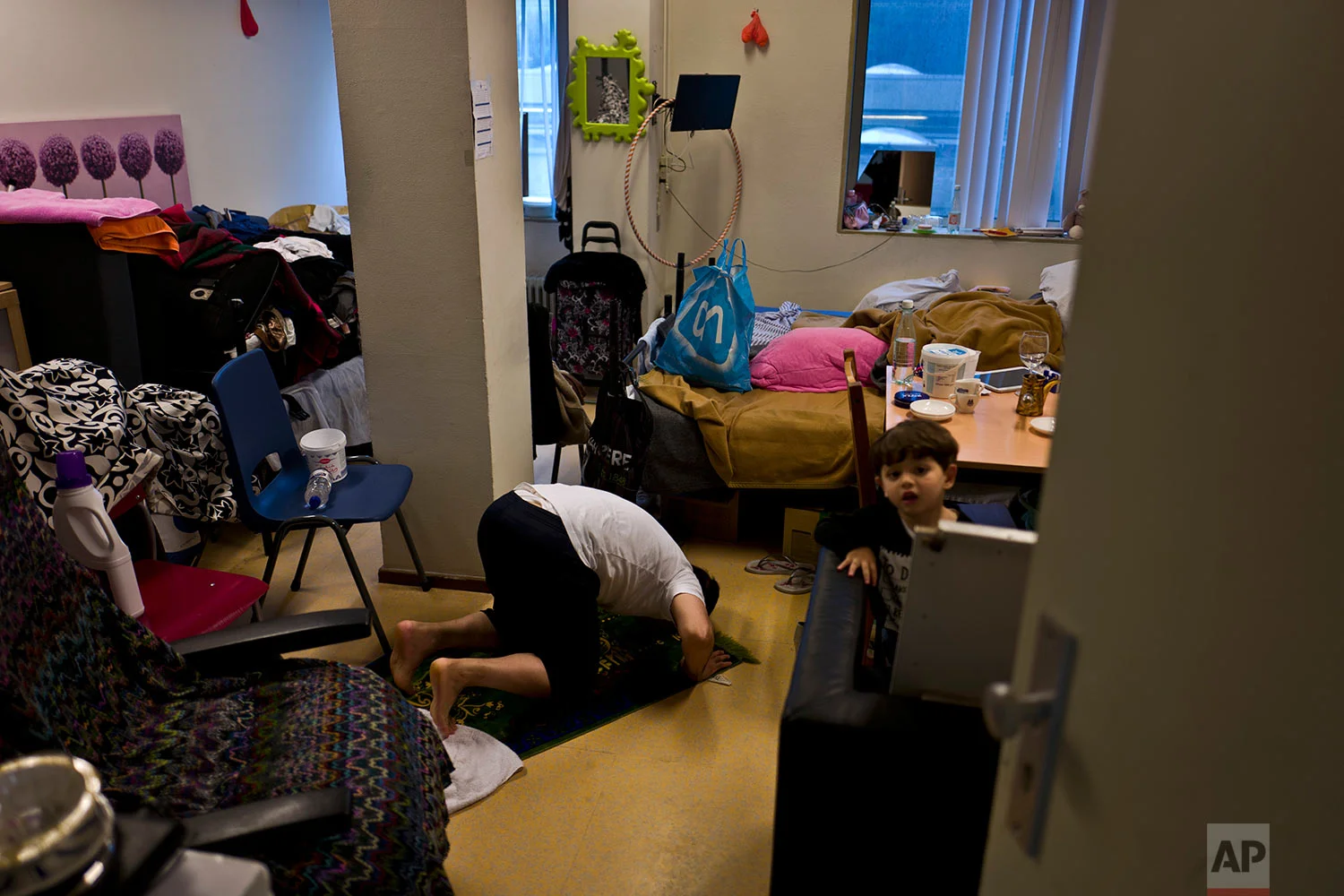 In this Wednesday, June 28, 2017 photo, refugee Hani Nashed, 53, from Aleppo, Syria, offers prayers while his young neighbour Lith Mashhadi, 3, a Syrian refugee from Aleppo, peeks on, at Hani's room in the former prison of Bijlmerbajes, in Amsterdam, Netherlands. (AP Photo/Muhammed Muheisen)
