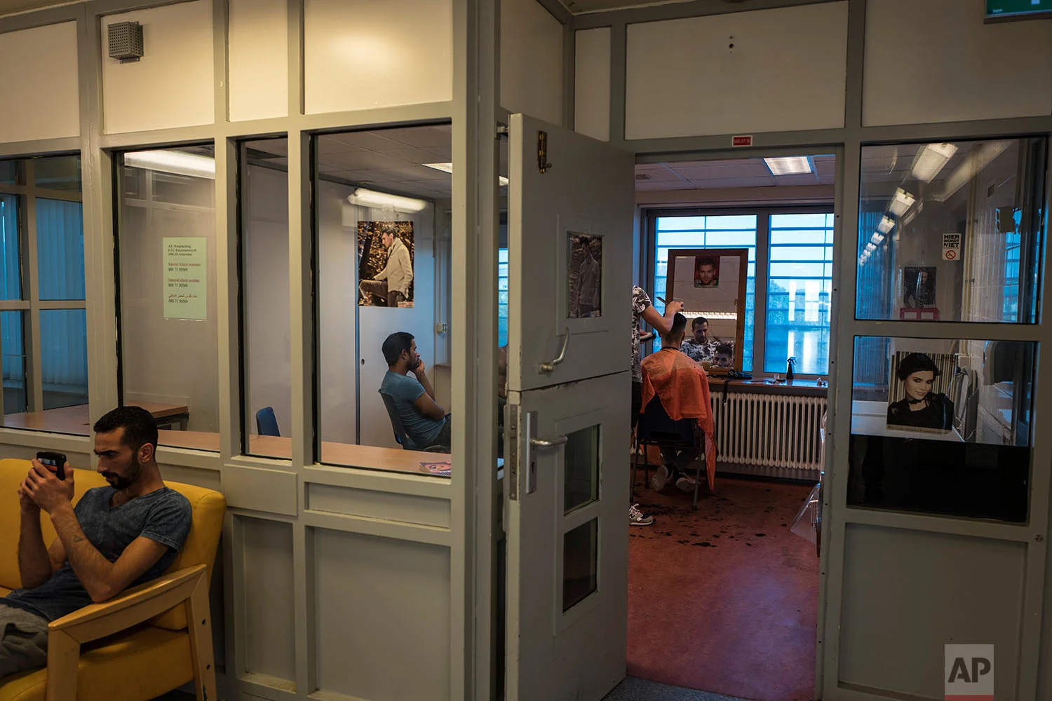 In this Friday, July 7, 2017 photo, Syrian refugee Ahmad Alshebly, 25, left, checks his mobile phone while Iranian migrant Hussein Azari, 25, is having a haircut by Iranian migrant Mohammed, 33, in a room at the former prison of Bijlmerbajes in Amsterdam, Netherlands. (AP Photo/Muhammed Muheisen)