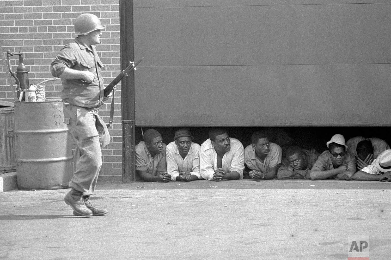 In this July 26, 1967 photo, an Army soldier stands guard as men captured in the vicinity of the 10th Police Precinct in Detroit peer from under a garage door awaiting transfer. The precinct building came under fire in daylight hours and an Army force, using armed personnel carriers and tanks came to the police station. (AP Photo)