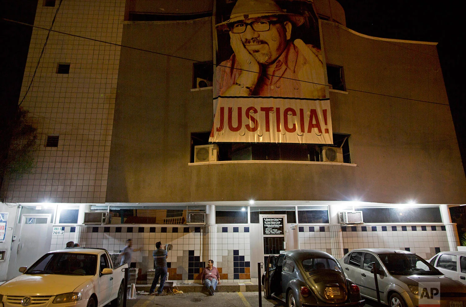 In this July 2, 2017 photo, a banner of slain journalist Javier Valdez and the word in Spanish "Justice!" hangs from the Riodoce newspaper building as drivers prepare to distribute the publication in Culiacan, Sinaloa state, Mexico. In the giant photo of Riodoce's fallen founder, his middle finger is displayed defiantly for all of Sinaloa to see. (AP Photo/Enric Marti)