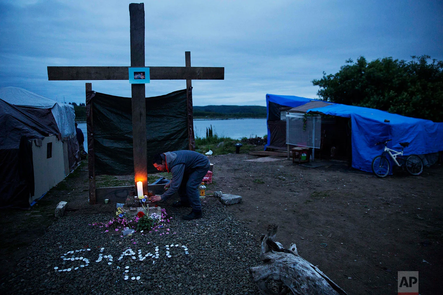 A mourner lights a candle at the memorial marking the spot where Shawn Vann Schreck died two days before in a homeless encampment where he lived along the river in Aberdeen, Wash., Wednesday June 14, 2017. On Aberdeen's banks, residents of the homeless encampment pulled driftwood from the water to construct a cross, 8 feet tall, to honor their latest loss: Vann Schreck, 42, who died slowly from heart and lung ailments made worse by infrequent medical care and longtime addiction. A generation ago, people like him went to work in the mills and bought tidy houses in nice neighborhoods, says the Rev. Sarah Monroe, a street minister here. (AP Photo/David Goldman)
