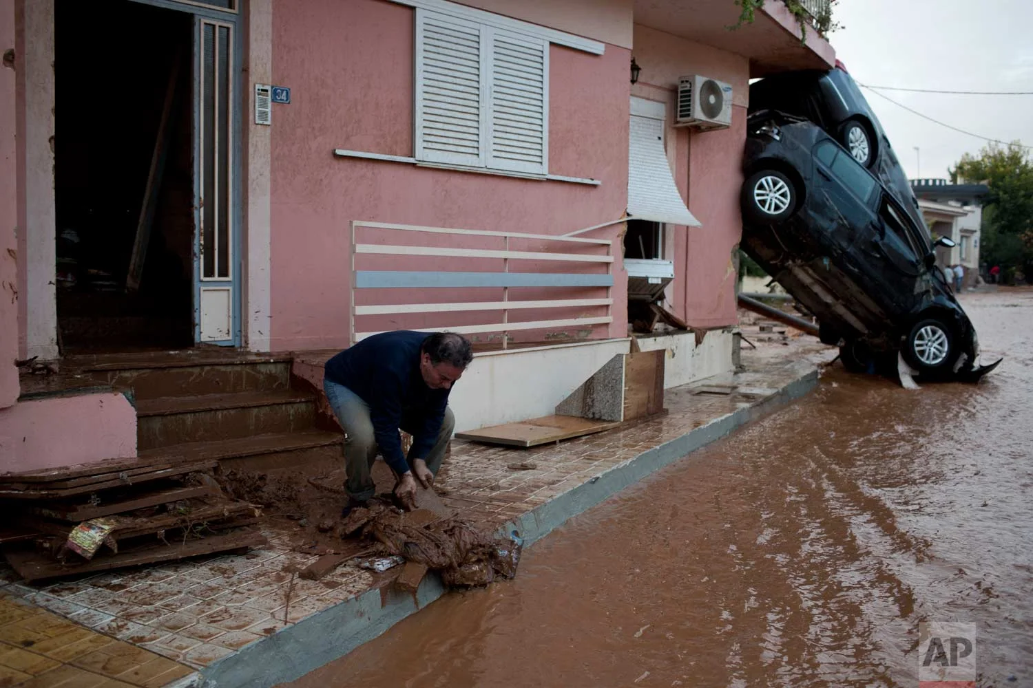 In this Wednesday, Nov. 15, 2017 photo, a man clears mad from his home in front of flipped over cars in the town of Mandra western Athens, following deadly flash floods that hit the area. Nearly 1,000 buildings were damaged, most of them private houses, as well as businesses, storage areas and public buildings.(AP Photo/Petros Giannakouris)