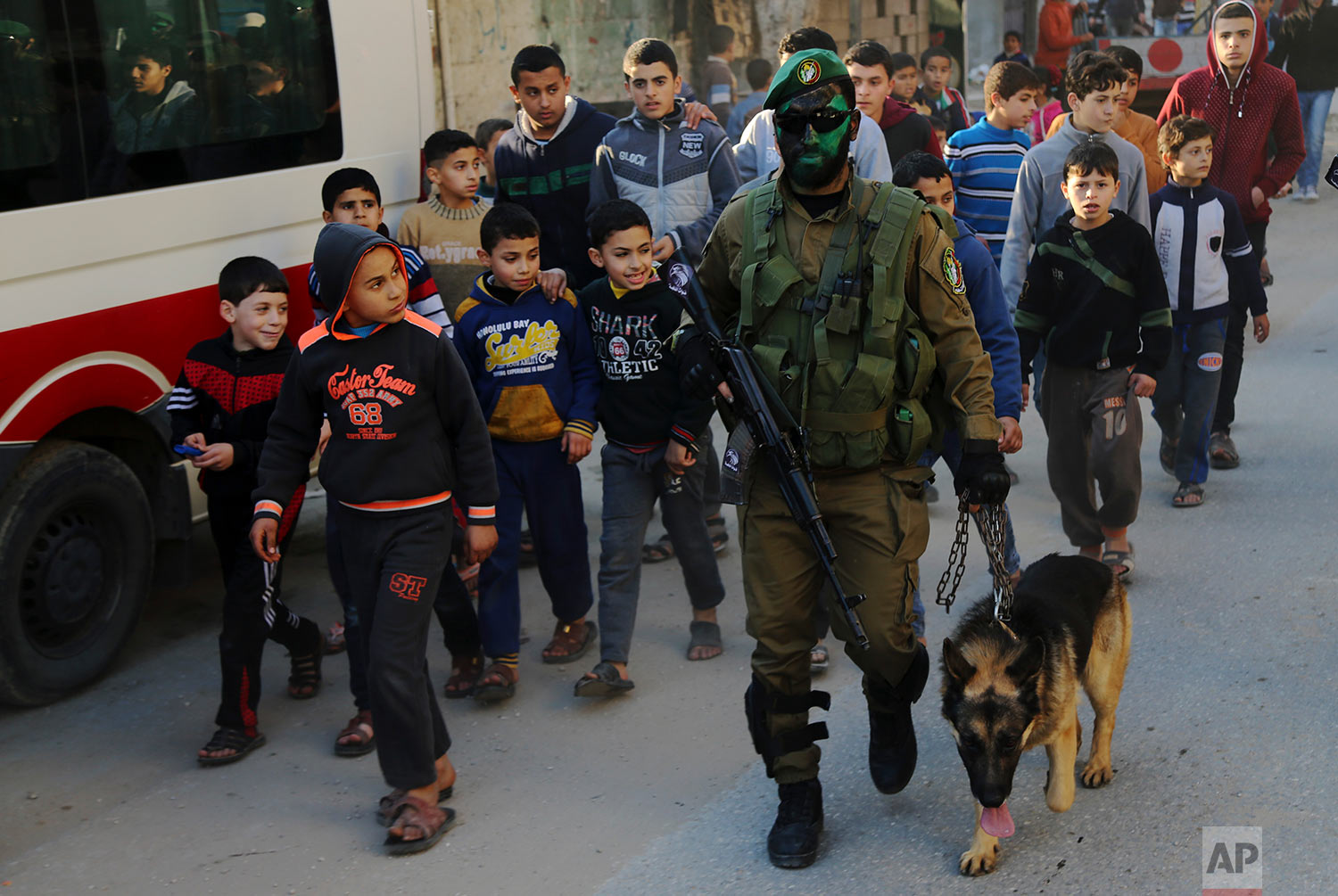 Palestinian youths watch a masked militant from the Izzedine al-Qassam Brigades, a military wing of Hamas, marching with a dog along the streets of Nusseirat refugee camp, Central Gaza Strip, Thursday, Jan. 19, 2017. Hamas armed wing has added a new dogs unit to its battalions. (AP Photo/Adel Hana)