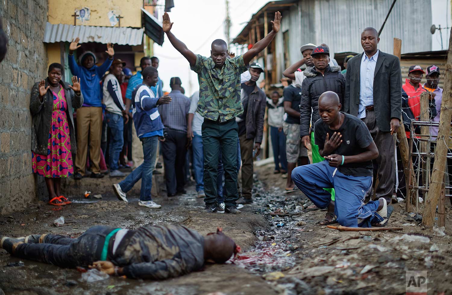  In this Wednesday, Aug. 9, 2017 photo, residents hold their hands up in the air towards police, as a man genuflects, right, next to the body of a man who had been shot in the head and who the crowd claimed had been shot by police, in the Mathare slu