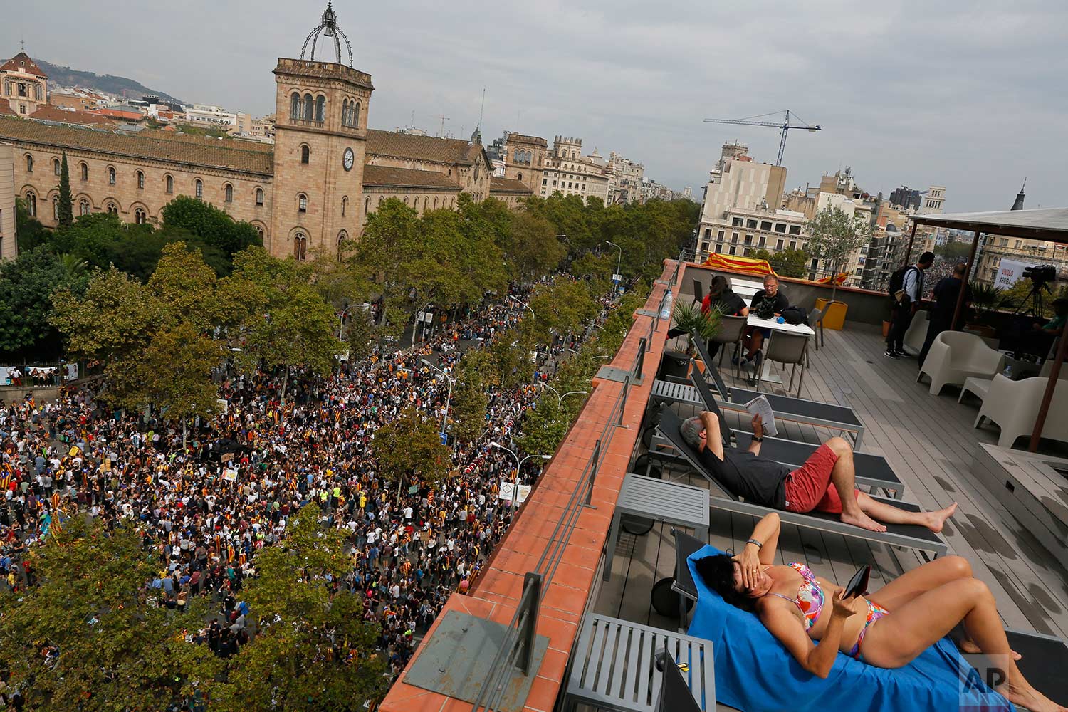  In this Tuesday Oct. 3, 2017 photo, a couple, tourists from Switzerland, sunbath at a terrace overlooking Universitat square as demonstrators gather in protest in downtown Barcelona, Spain. Labor unions and grassroots pro-independence groups are urg
