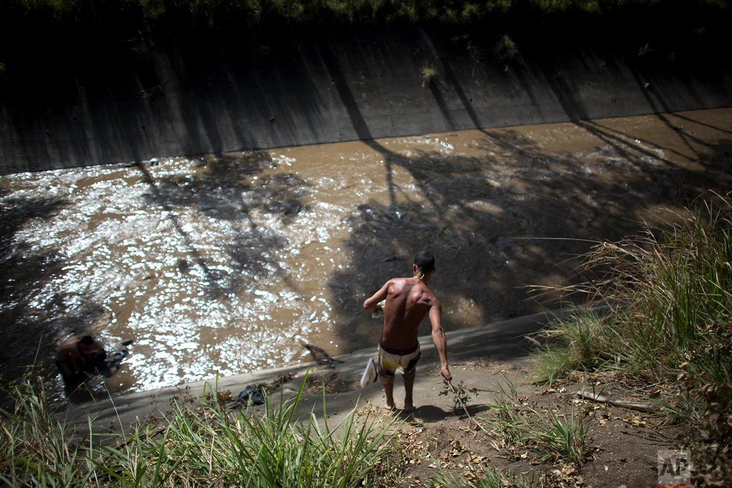 In this Dec. 5, 2017 photo, a man holds on to a branch to lower himself down the concrete bank to the polluted Guaire River where he will search for anything valuable he can sell, in Caracas, Venezuela. Most of the scavengers stream down from hillside barrios, and from afar, they seem to play in the river, but in reality their desperation to earn a living is what pushes them to risk entering the toxic, sewage water. (AP Photo/Ariana Cubillos)