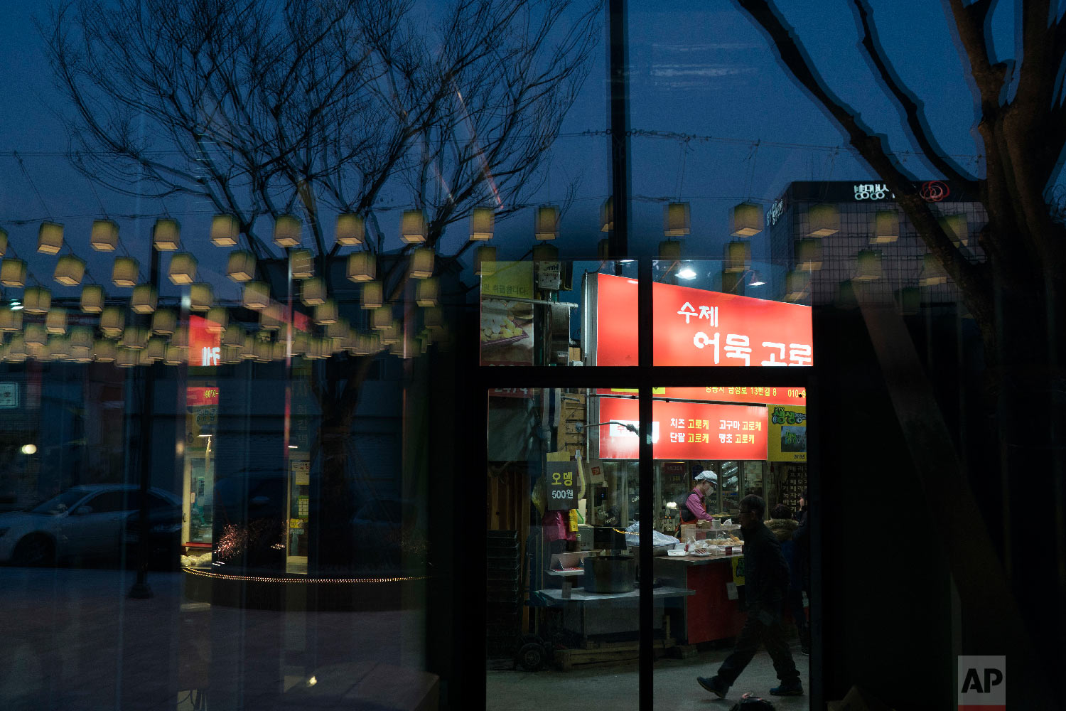 Decorative lamps are reflected on a glass door as a man walks past a restaurant at the traditional market in Gangneung, South Korea, Tuesday, Feb. 13, 2018. Markets in South Korea can range in size from simple gatherings held every five days or so in some rural areas to the massive Namdaemun market area in Seoul, which houses more than 10,000 stores selling anything from fur coats to sneakers to deep-fried shrimp balls. (AP Photo/Felipe Dana)