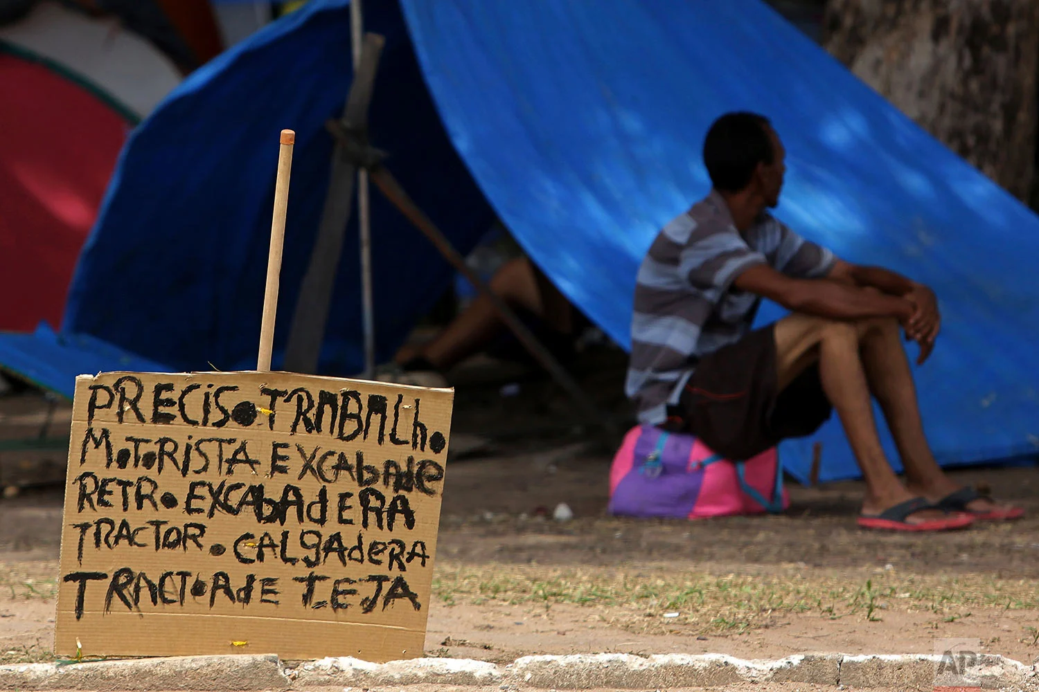 A sign that reads in Portuguese: "I need work. Tractor driver. Excavator." stands near a Venezuelan migrant outside his tent in Simon Bolivar Square where many Venezuelans have set up tents in Boa Vista, Roraima, Brazil, March 11, 2018. (AP Photo/Eraldo Peres)
