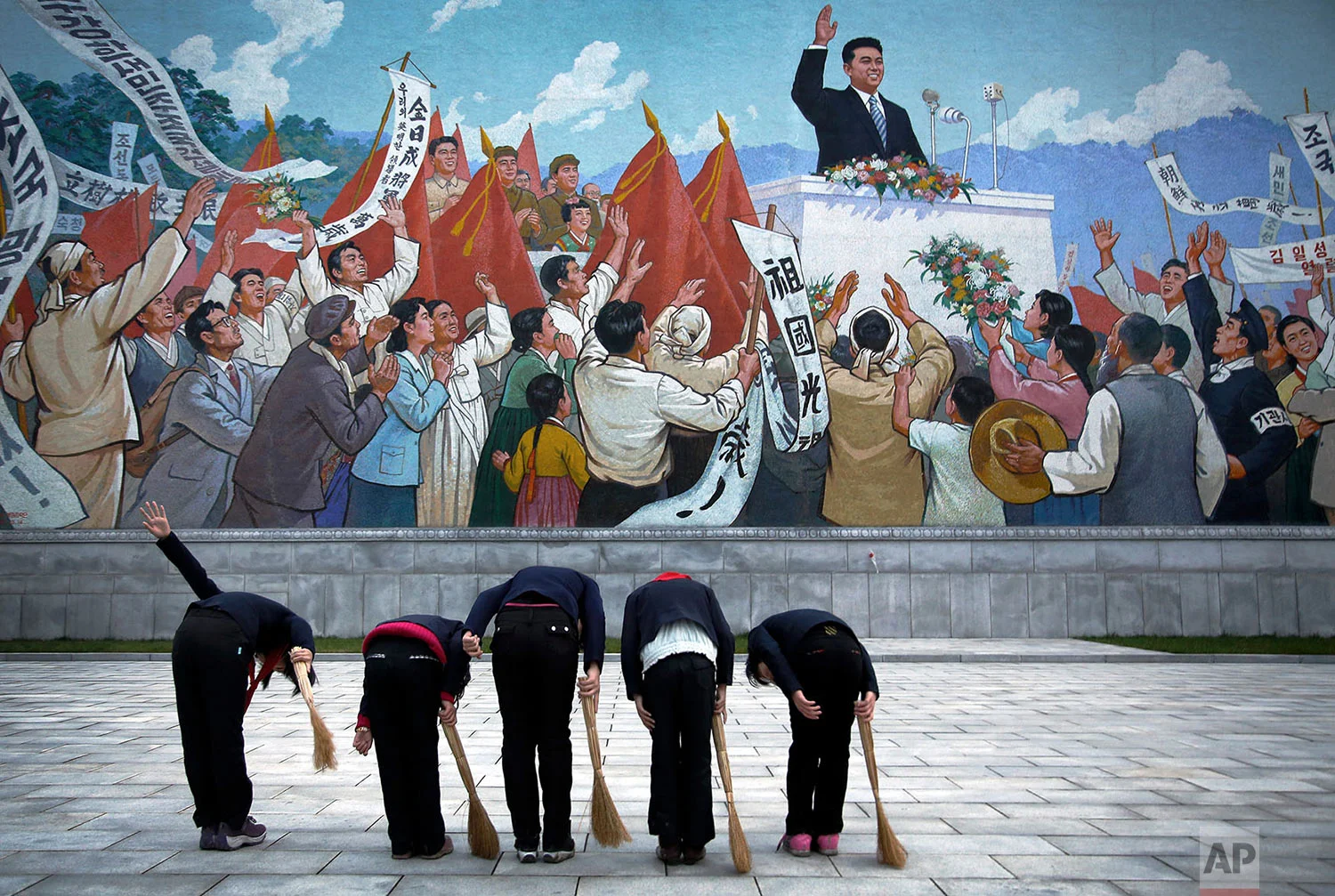 School girls holding brooms bow to pay their respects toward a mural that depicts the late North Korean leader Kim Il Sung delivering a speech, before sweeping the area surrounding the mural on Dec. 1, 2015, in Pyongyang, North Korea. (AP Photo/Wong Maye-E)